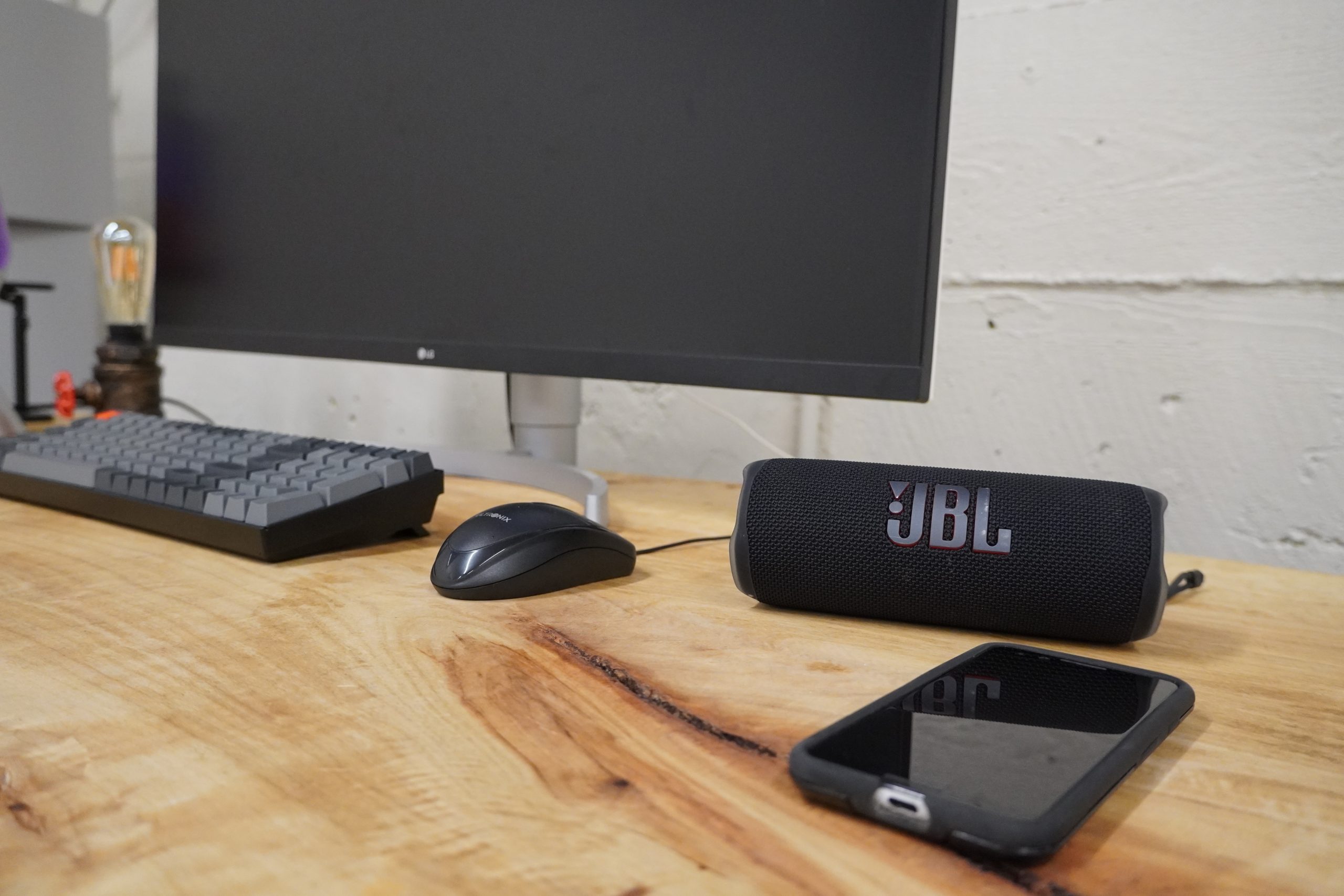 JBL Flip 6 On Desk Bluetooth Multipoint A JBL Flip 6 Bluetooth speaker sitting on a wooden desk next to a smartphone at its right and a monitor, keyboard, and mouse to its left.